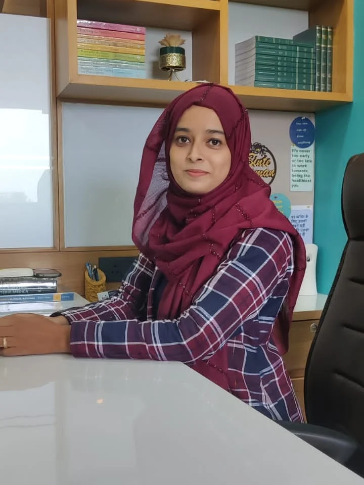 Dr. Gulfisha Maru seated at her consulting desk, the clinic's turquoise wall behind her